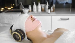 Image of Young woman on a salon bed with a pink alginate mask on her face, with headphones over her ears.